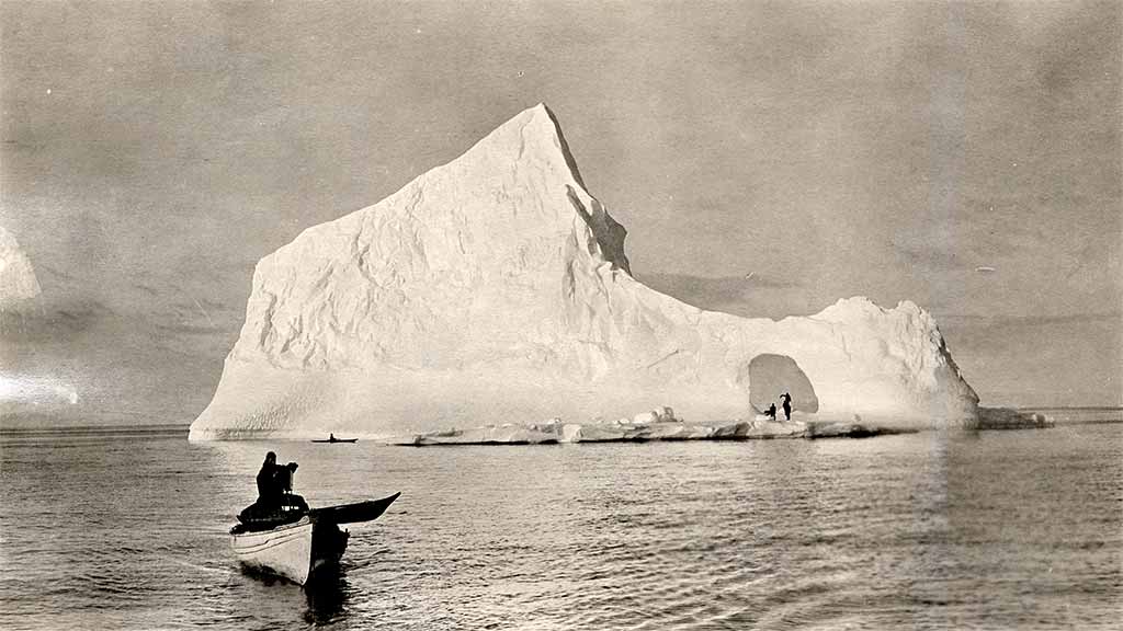 researchers in boat photograph distant iceberg with people under an arch