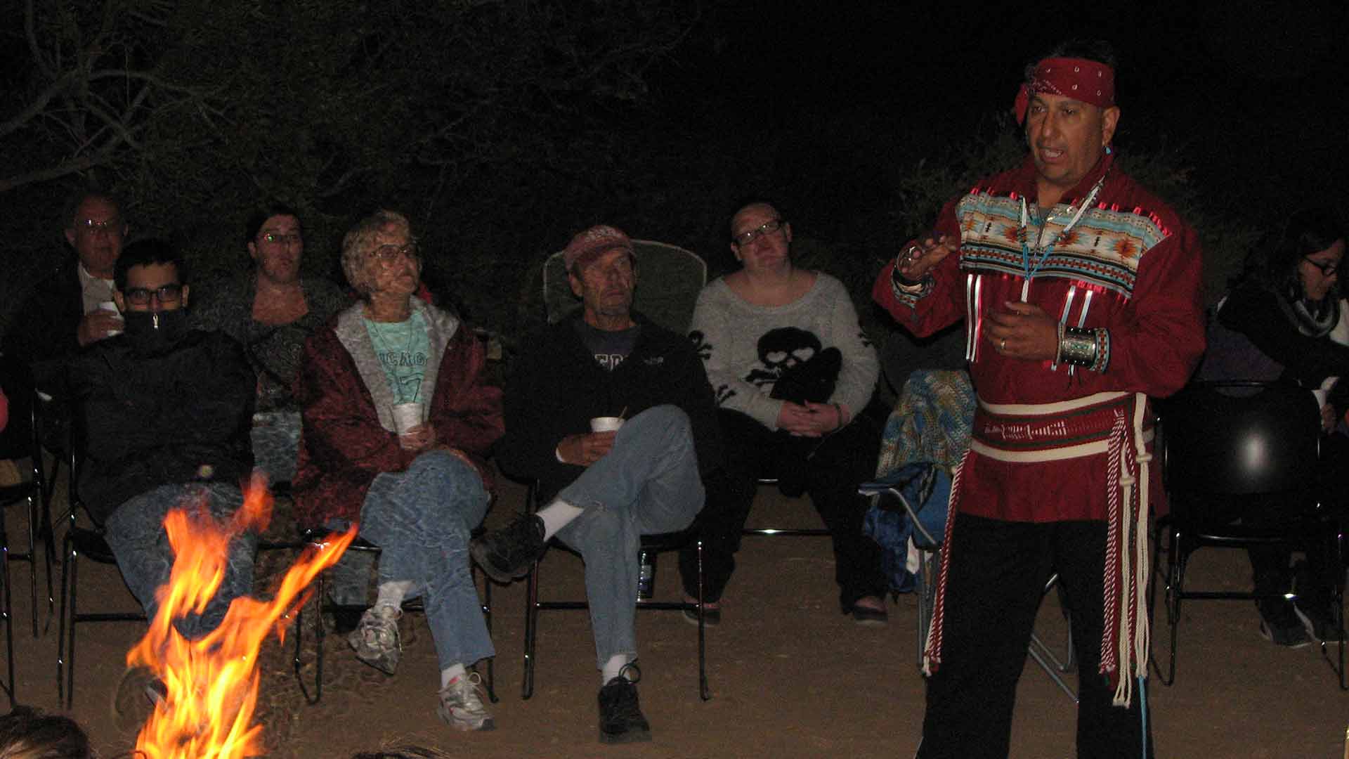 Alex Mares speaks to a seated group around an evening campfire