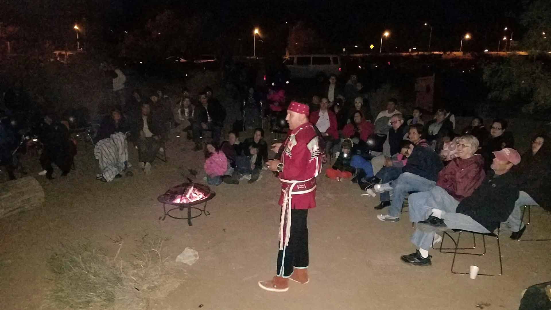 Alex Mares stands in front of a fire pit surrounded by people seeted in camping chairs with coats and blankets