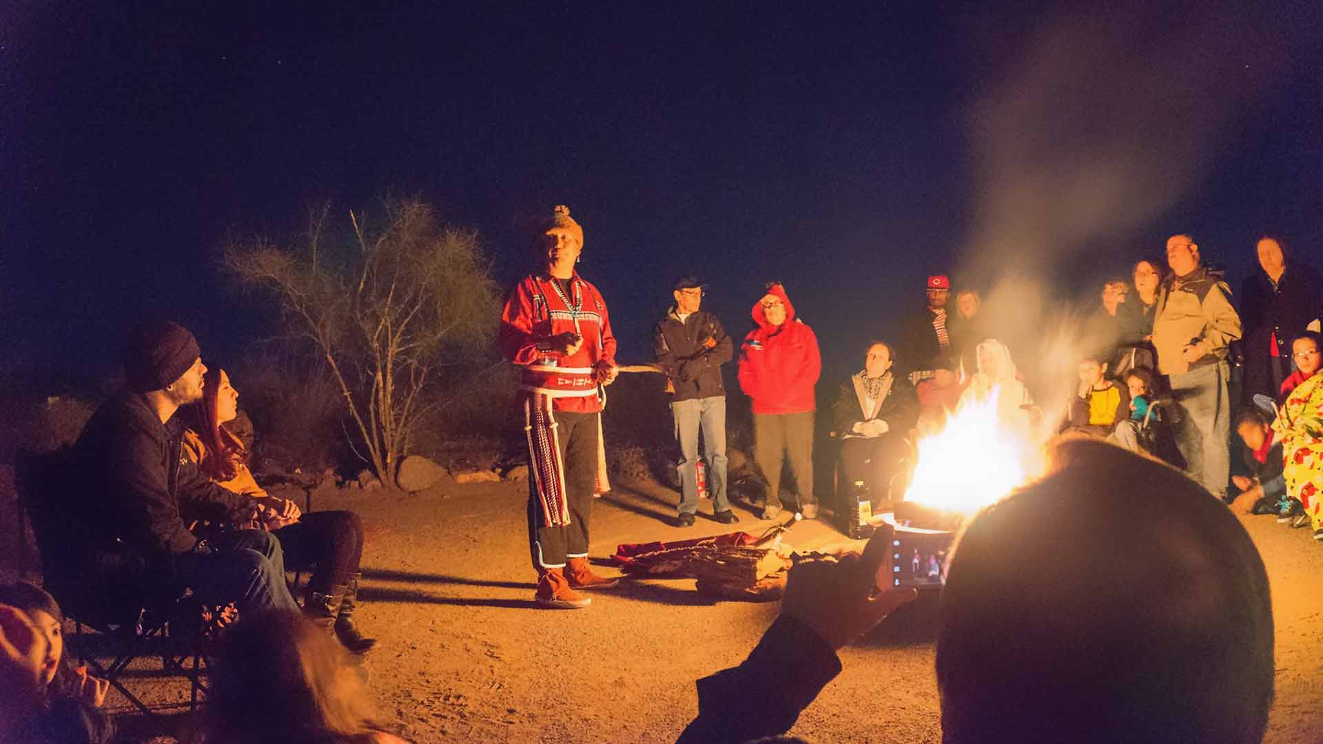 Alex Mares stands at a bright glowing campfire surrounded by people listening