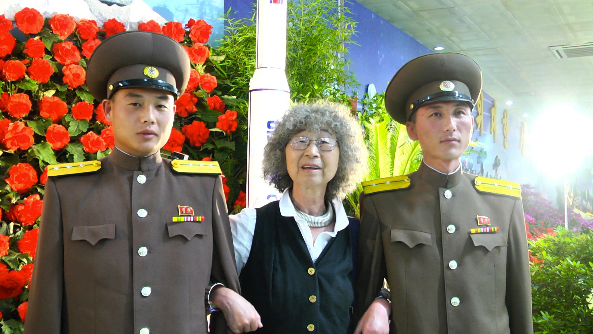 Korean military men arm in arm with elderly Korean woman in front of flowers and model rocket
