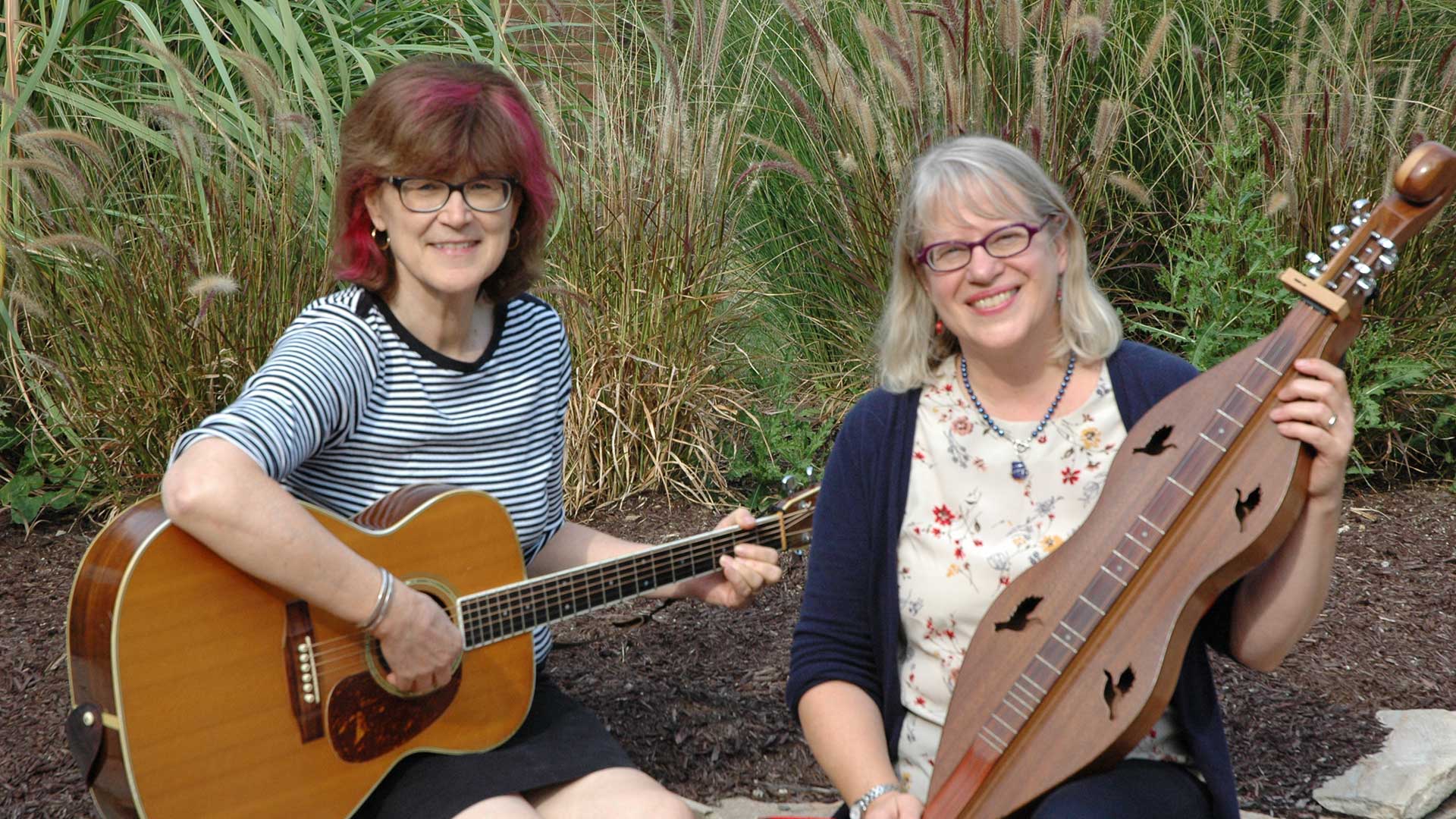 Two women sit in front of prairie grass, holding a guitar and dulcimer