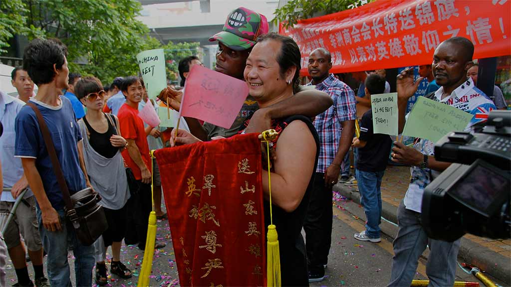 Chinese and African people intermingle and pose for a photo in the street with signs in Chinese and a video camera