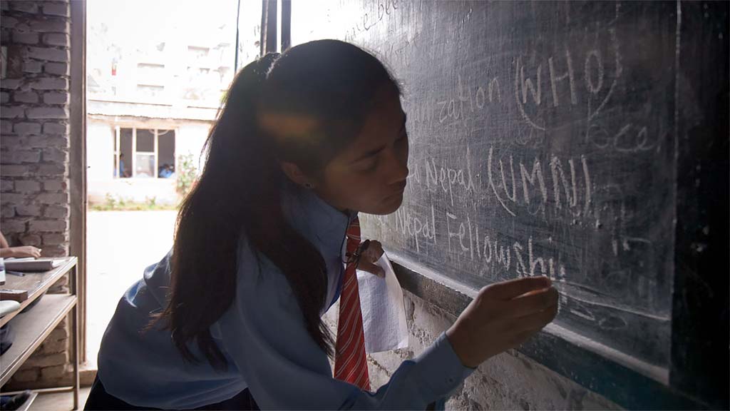 A female student writes "Nepal Fellowship" on a blackboard in a darkened classroom as daylight streams in