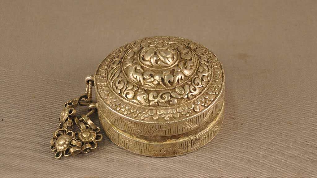 An ornate, round, silver container with a lid connected to the base by a decorative chain.