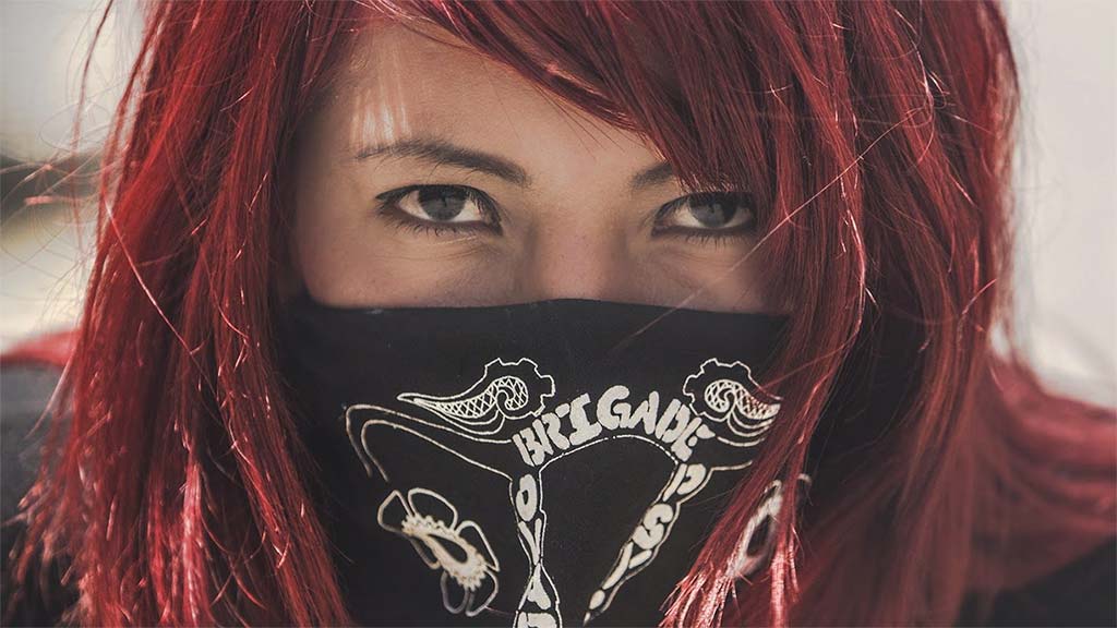 Woman with red hair wearing a bandana over her mouth looking forward