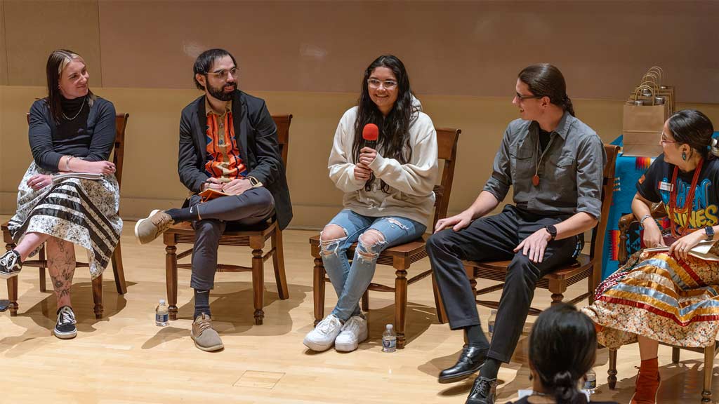 a panel of five individuals smile and chat with microphone on a stage