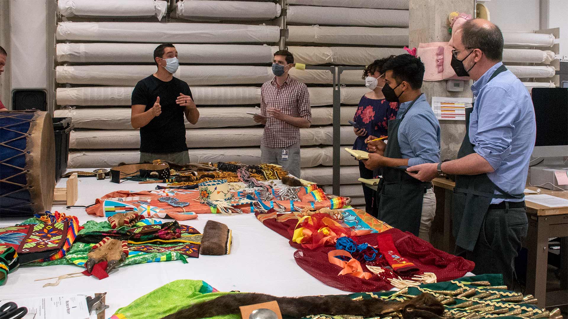 people in clipboards gather around a table of multicolored textiles as a individual leads a discussion