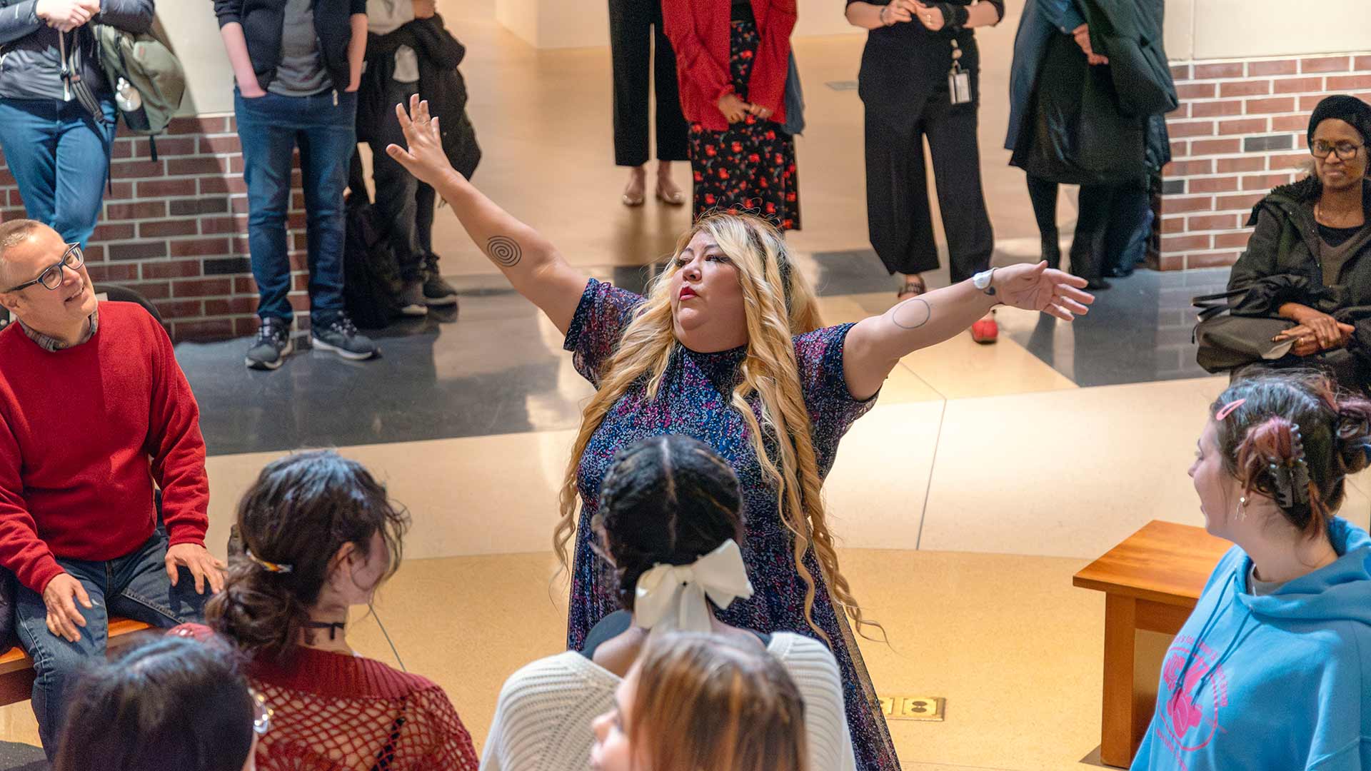 a woman in a gathering space with arms outstretched as audience members look on from around the room