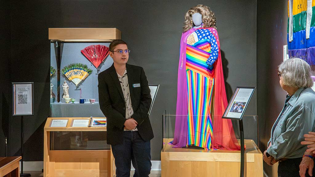 a staff member leads a tour in an exhibit space in front of a colorful costume and case of artifacts