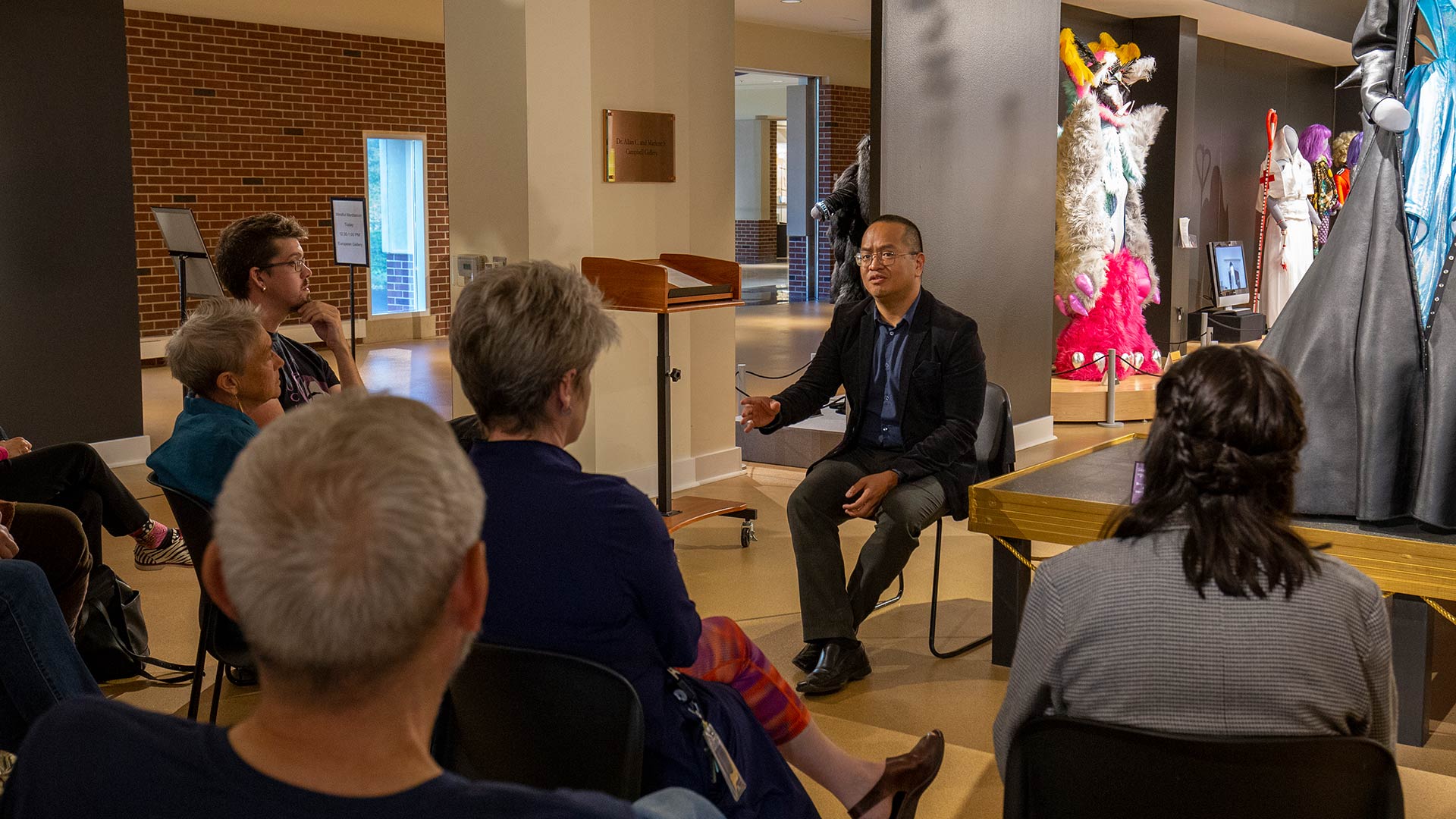 a speaker in conversation with a group of visitors in front of drag costumes