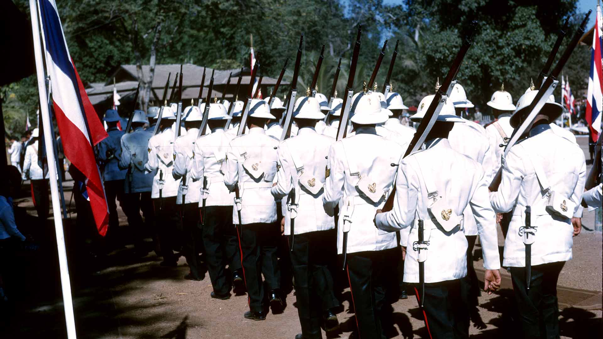 A procession of about 20  guards in white military dress and weaponry