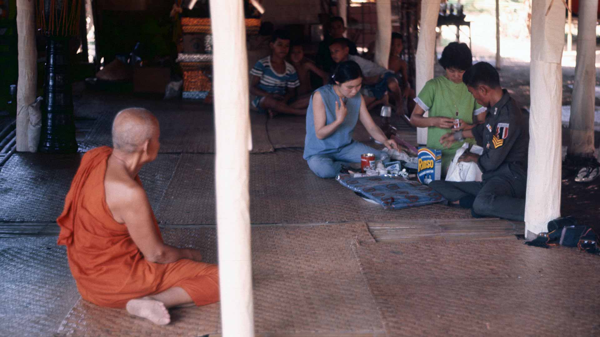 A seated monk looks on as a trio of people prepare gifts on the other side of the room