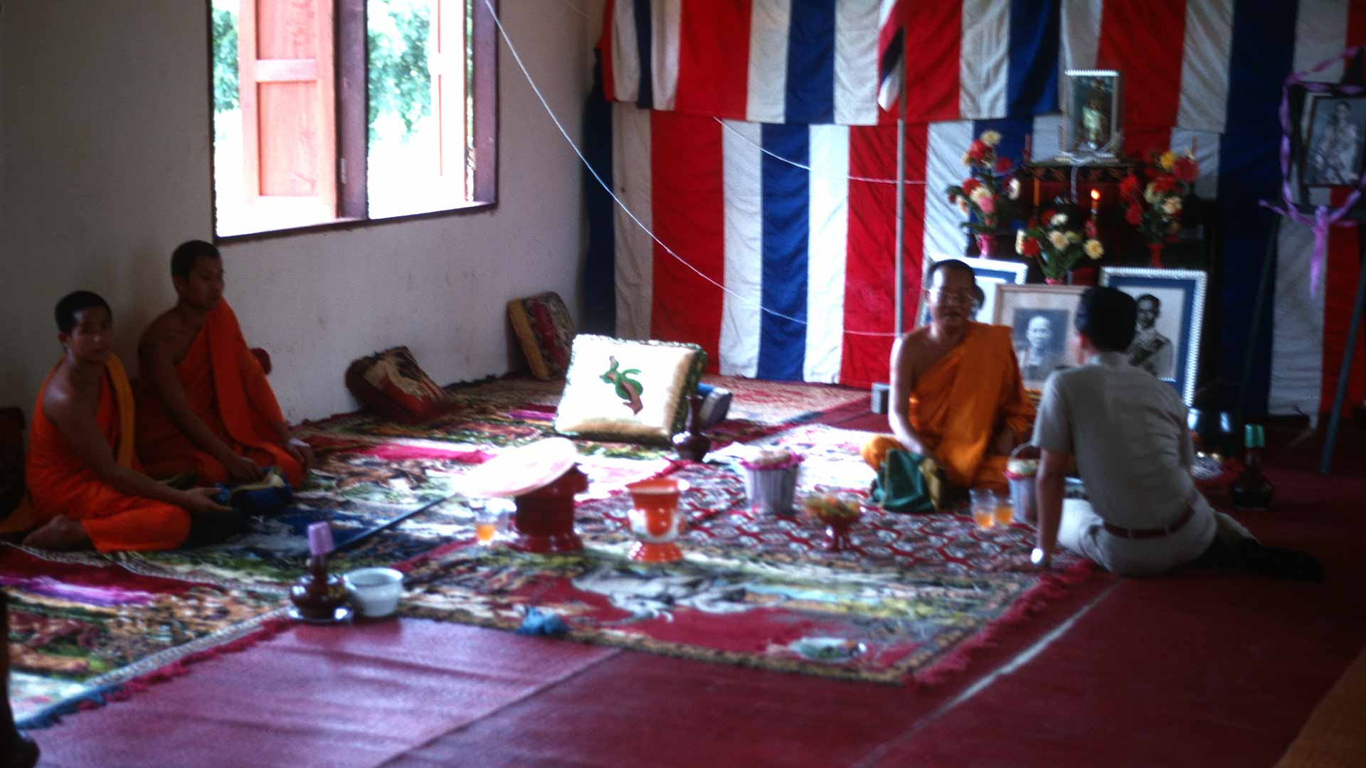 Monk receiving visitors in visiting area sitting on floor with thai flags cover back wall