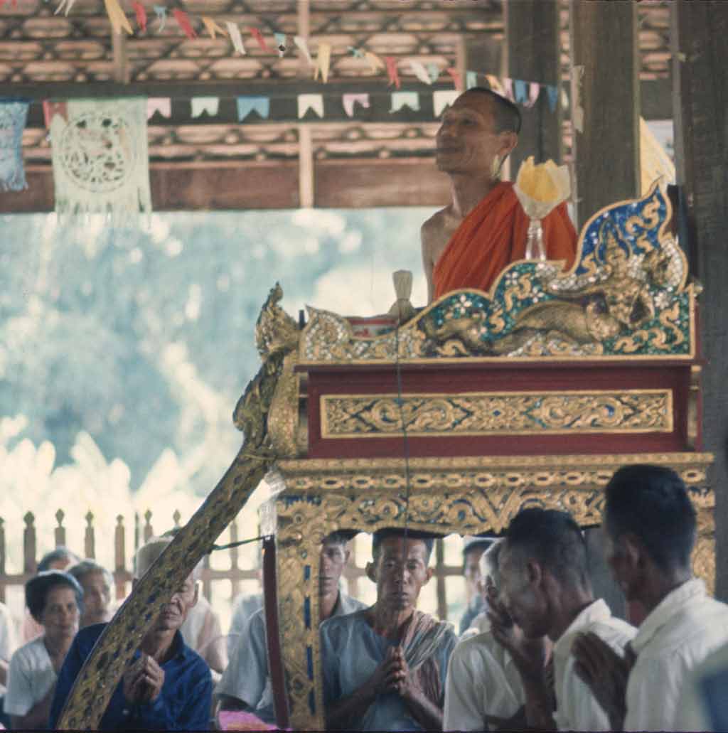 A man on a decorated, raised platform. Villagers are seated below him with their palms pressed together.