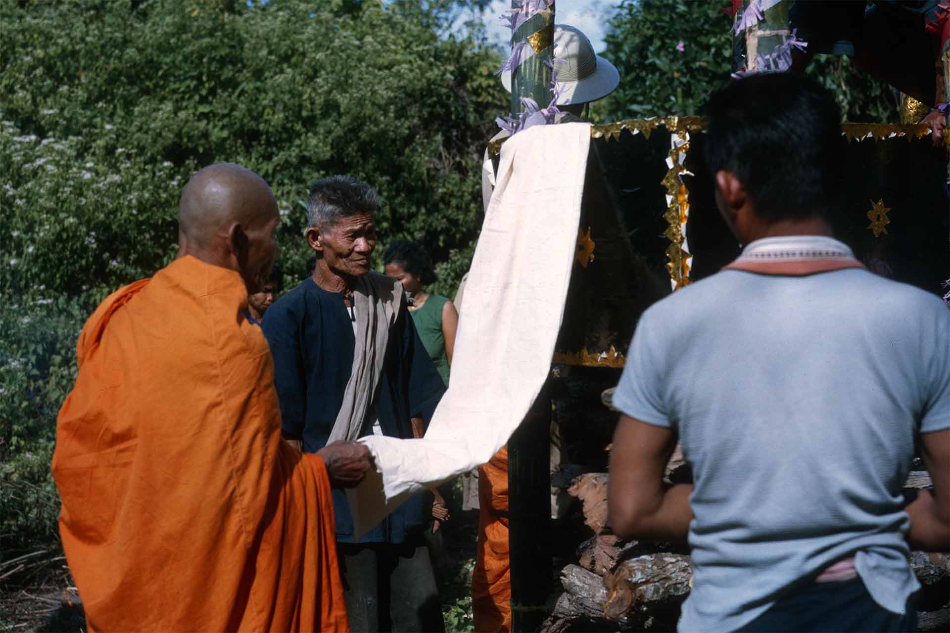 monk holding cloth attached to casket, people surround funeral pyre