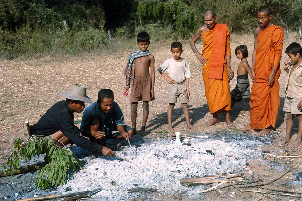 man sifting through ash for bones, monks and kids gathered watching