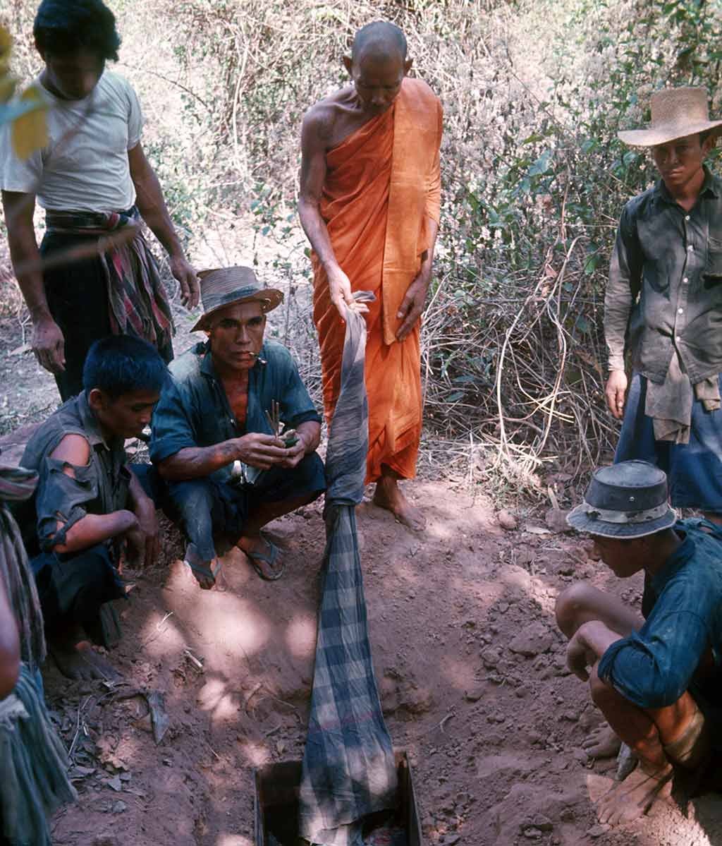 monk holding cloth staring down, in clearing, people around him