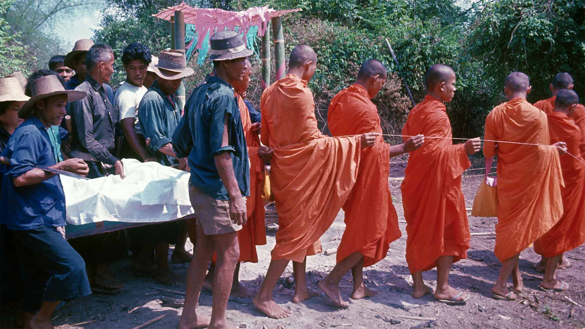 on left side villagers surround coffin draped in white cloth, to the right procession of monks holding string move outward