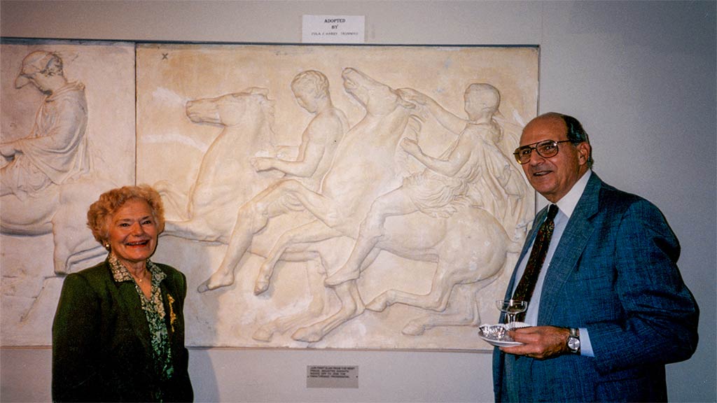 Pola and husband stand in front of the parthenon frieze panel they adopted at a musuem reception