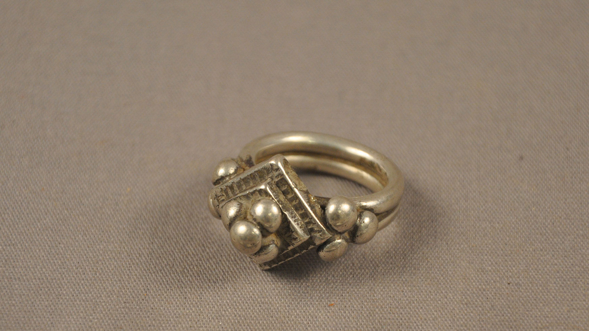 A silver-colored metallic ring with an indentation running through its band. It has an attachment consisting of two flat, silver-colored squares with four circular silver colored objects on top of the smaller square. On both of the sides of the squares are three circular silver colored objects.