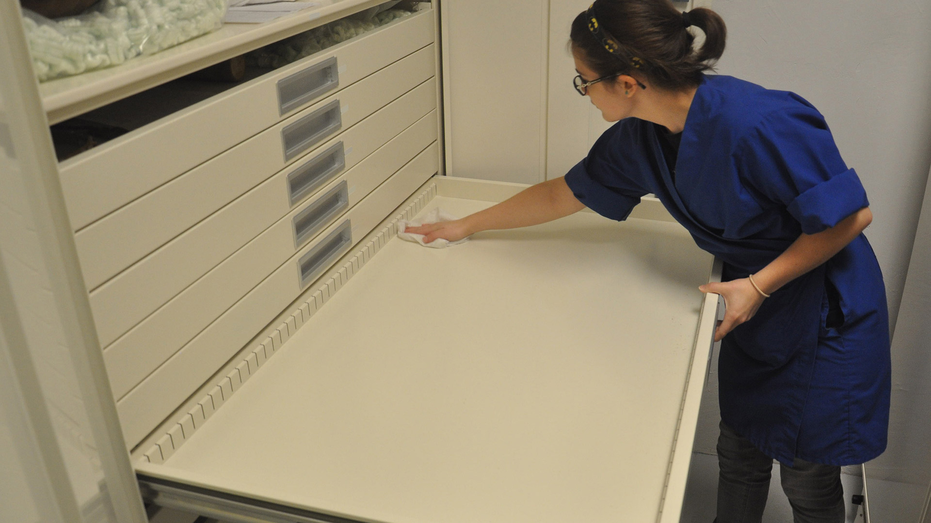 A woman in a blue smock cleans a shallow, empty metal drawer using a washrag