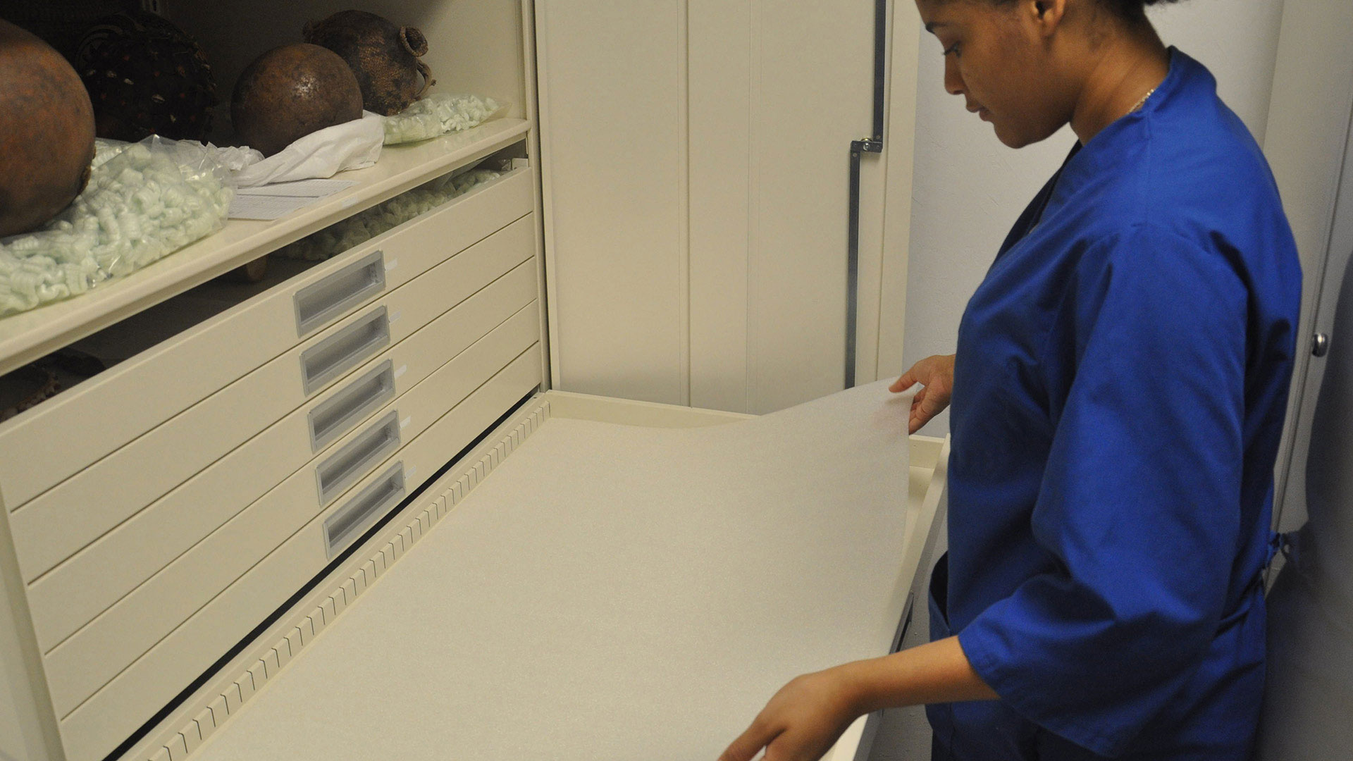 A woman in a blue smock lays a thin sheet of white-colored foam down within an opened drawer.