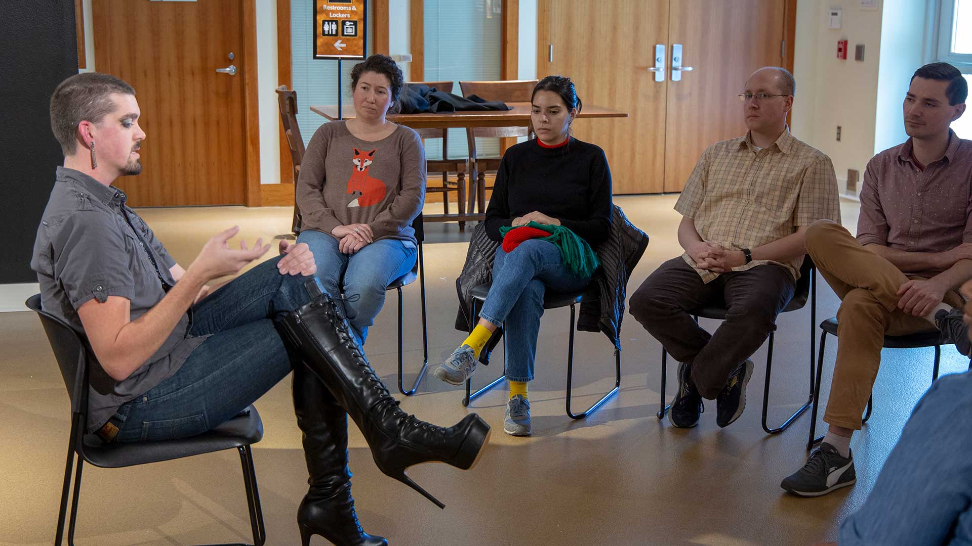 A seated man in makeup with earrings and stilletto platform boots speaks to several people in a museum gallery