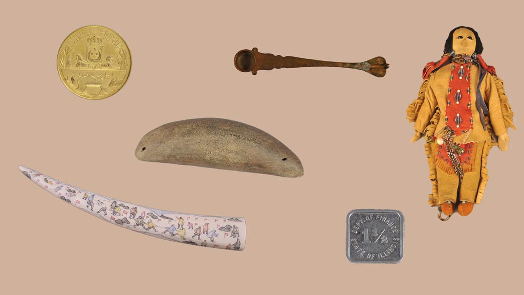 a gold medal, a curved carving with paintings on it, a curved wooden block, a small brass spoon, a small square silver token, and a doll with leather clothes and beadwork throughout