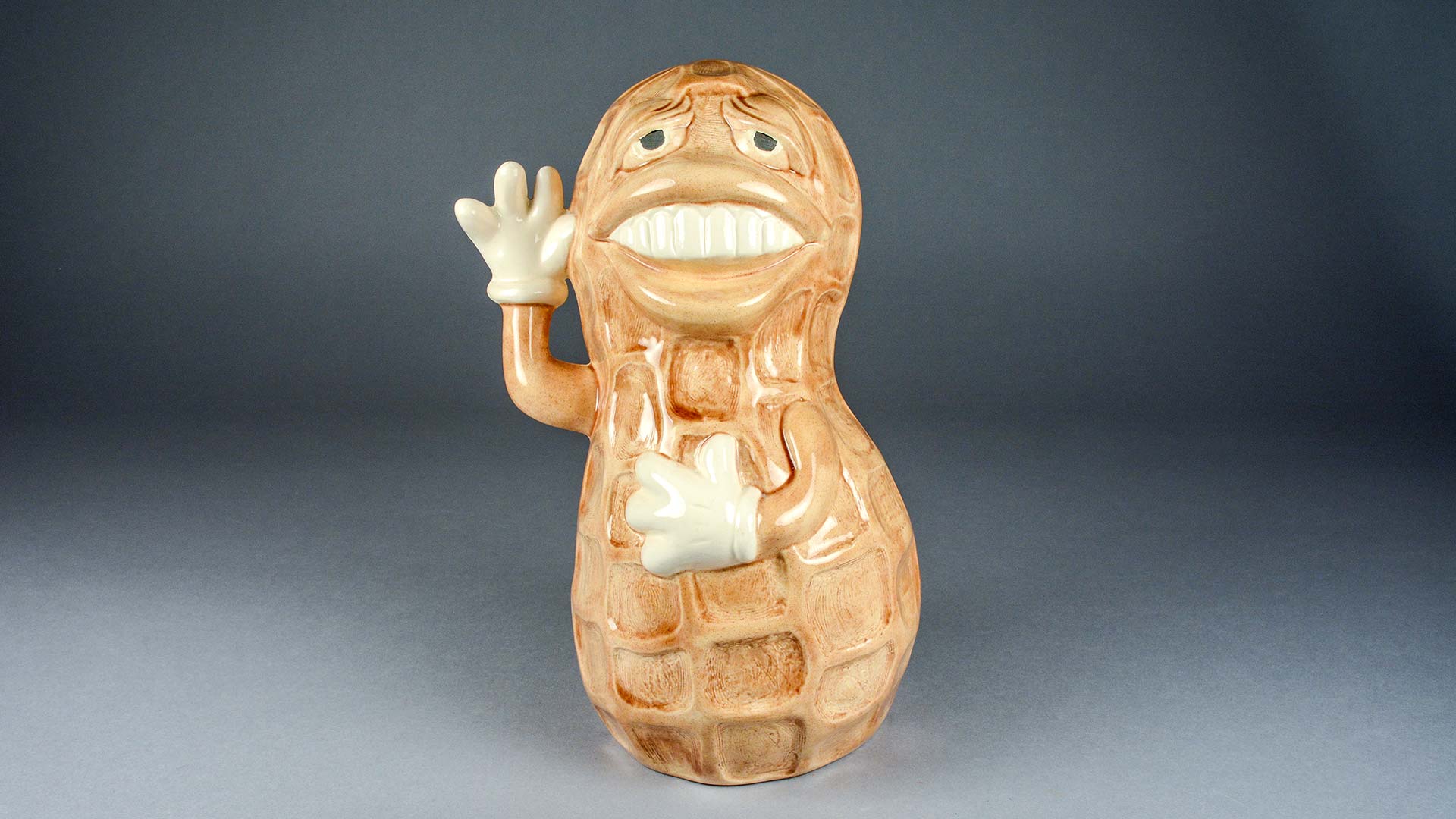 ceramic peanut sculpture with a comical smiling face, large lips, and a waving hand