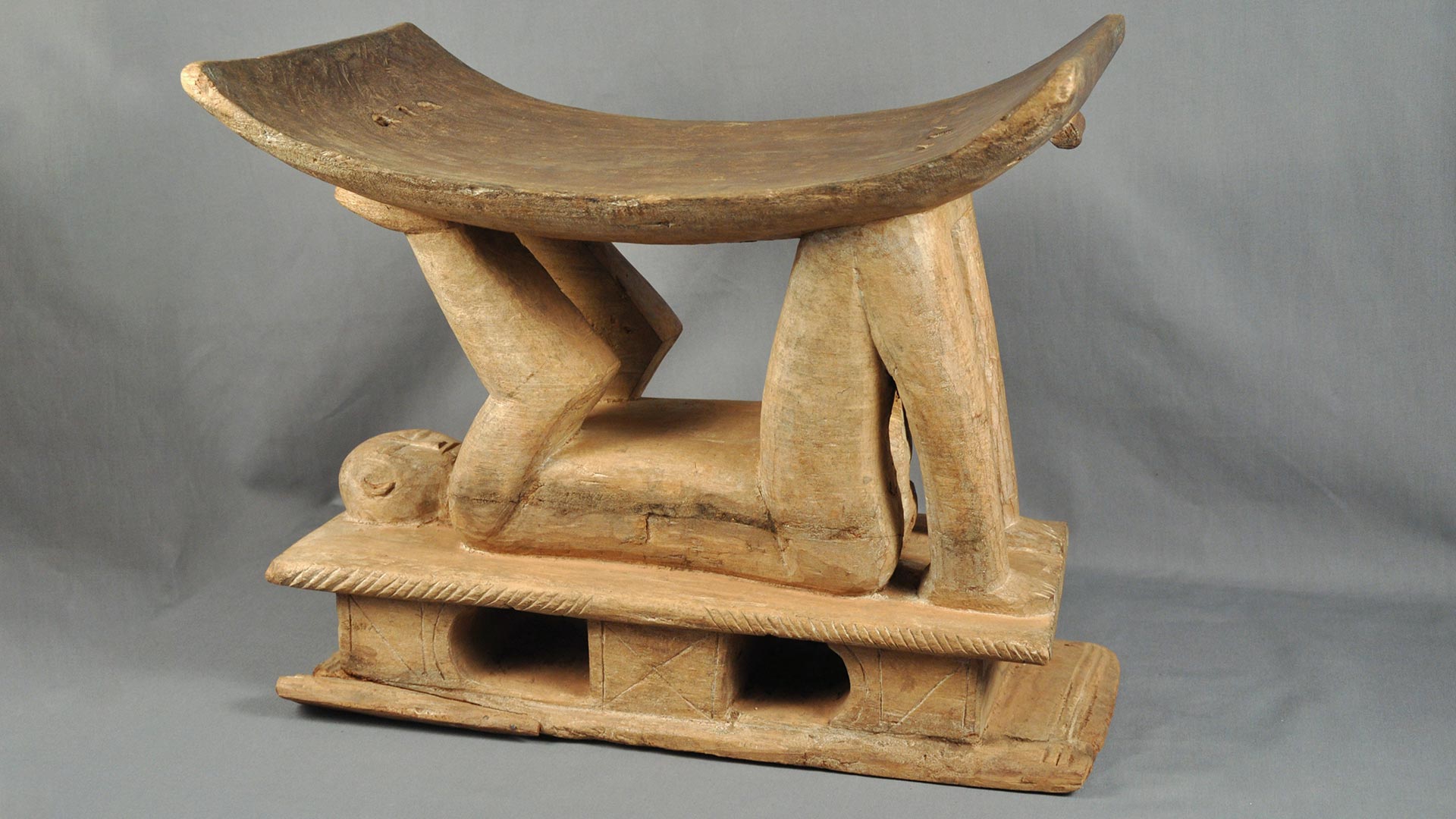 carved wooden stool with a carved human figure laying on their back, holding up a curved seat with their arms and knees