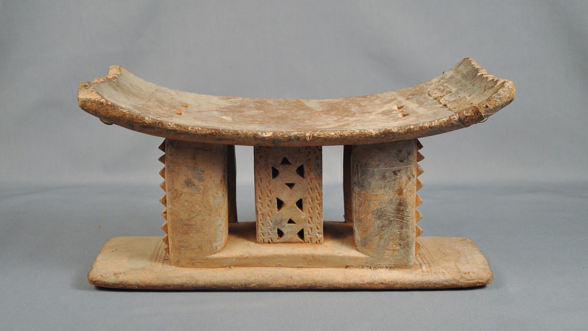 A wooden stool with a concave seat, and three carved pillared supports on a flat base