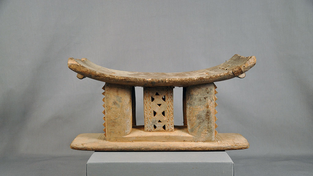 A wooden stool with a concave seat, and three carved pillared supports on a flat base