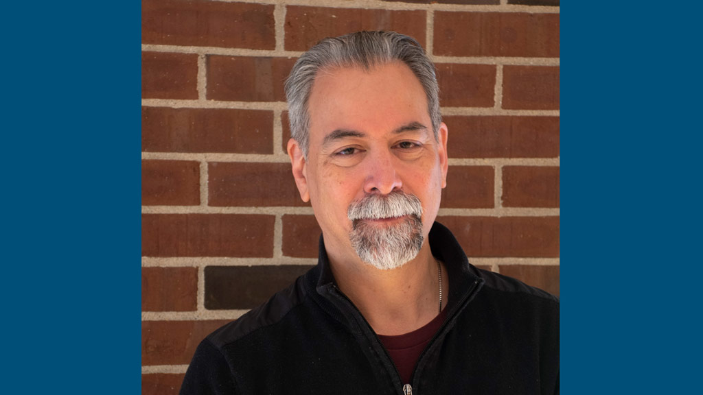 man with gray hair and beard standing against a brick wall, wearing a black jacket