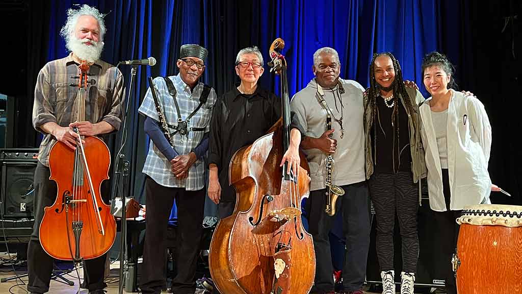 Six members of the MIYUMI Project ensemble pose on stage for a picture with stringed and reed instruments and a japanese drum