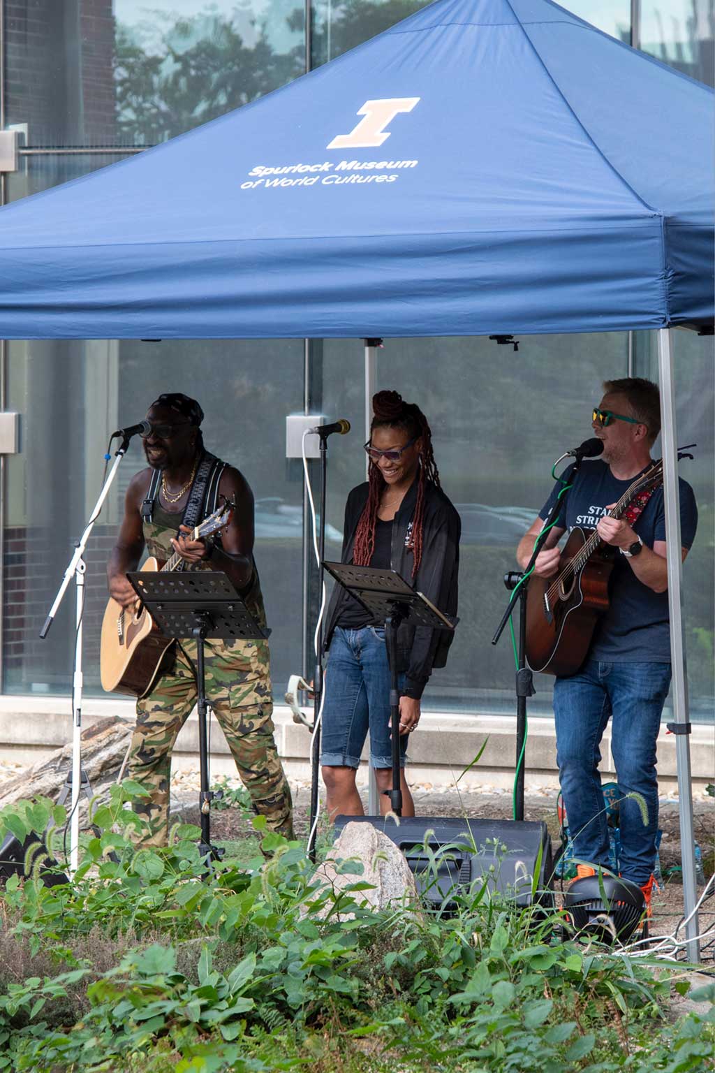 A vocalist and two guitarists perform under a tent outdoors in front of the museum