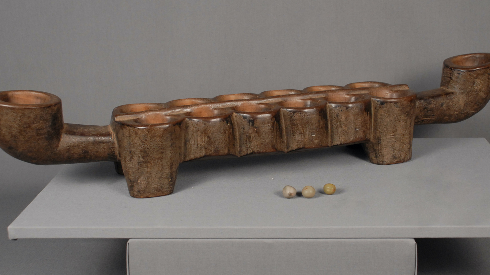 Wooden Mancala set with 12 holes in the middle and two holes on each side and three pebbles lying next to it.