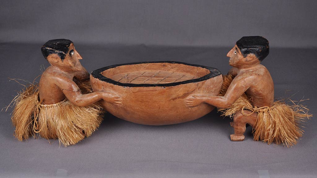 two carved male figures in grass skirts hold an oversized wooden bowl