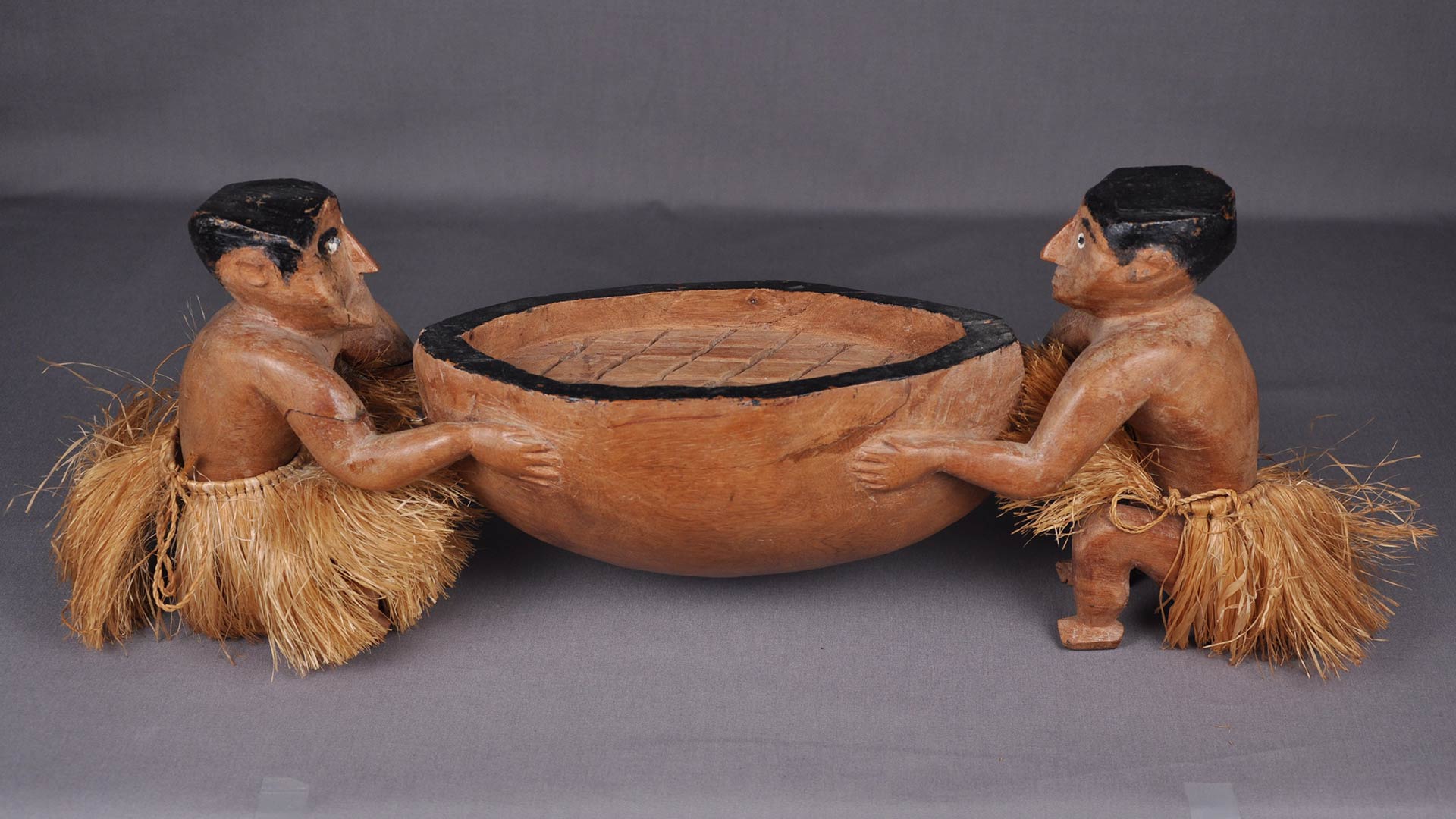 two carved male figures in grass skirts hold an oversized wooden bowl