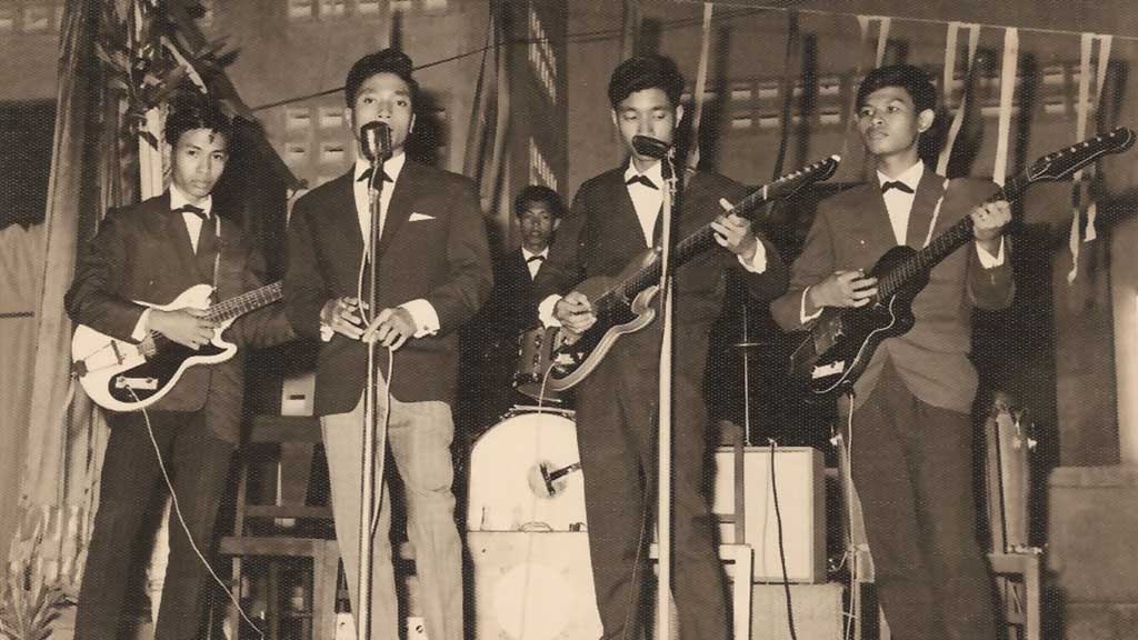 An old photo of four young bandmembers standing on stage in front of microphones