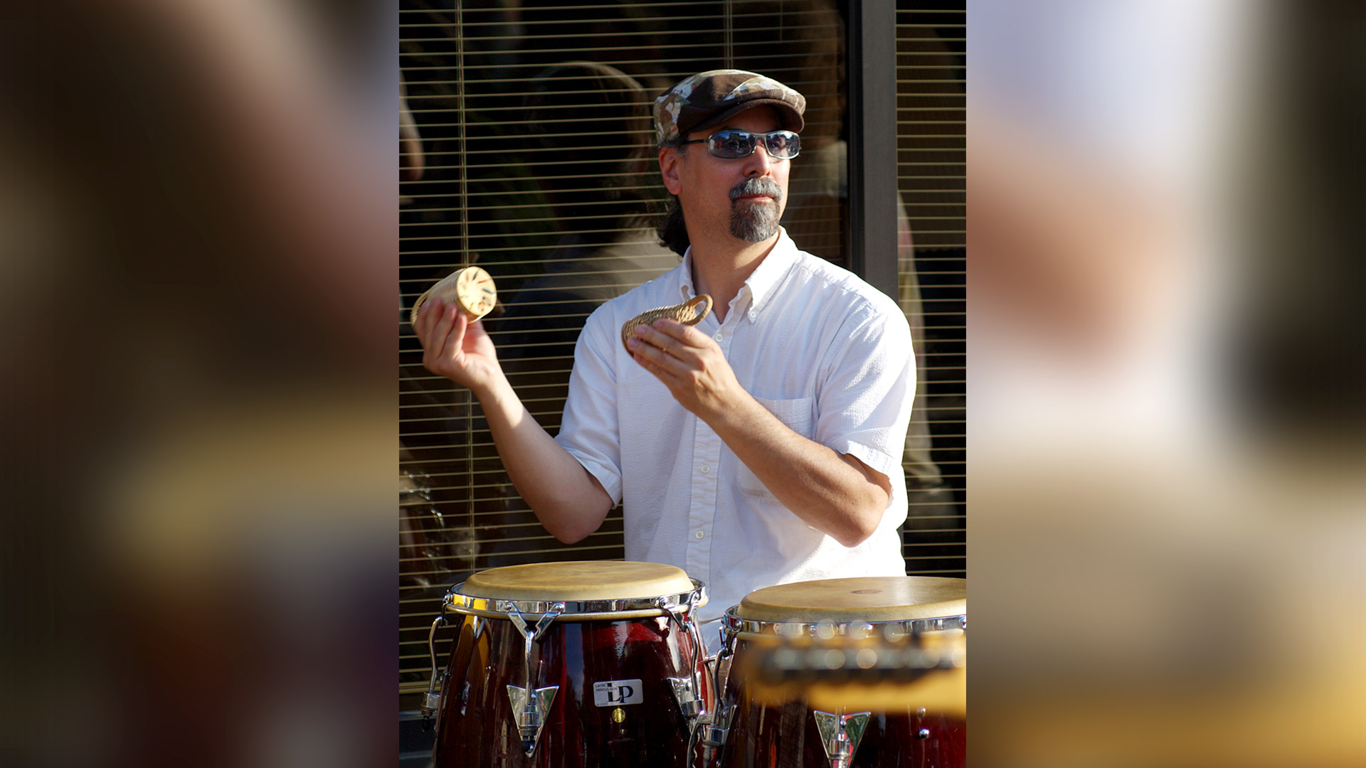 Man in button-down, sunglasses, and ascot hat playing shakers and drums