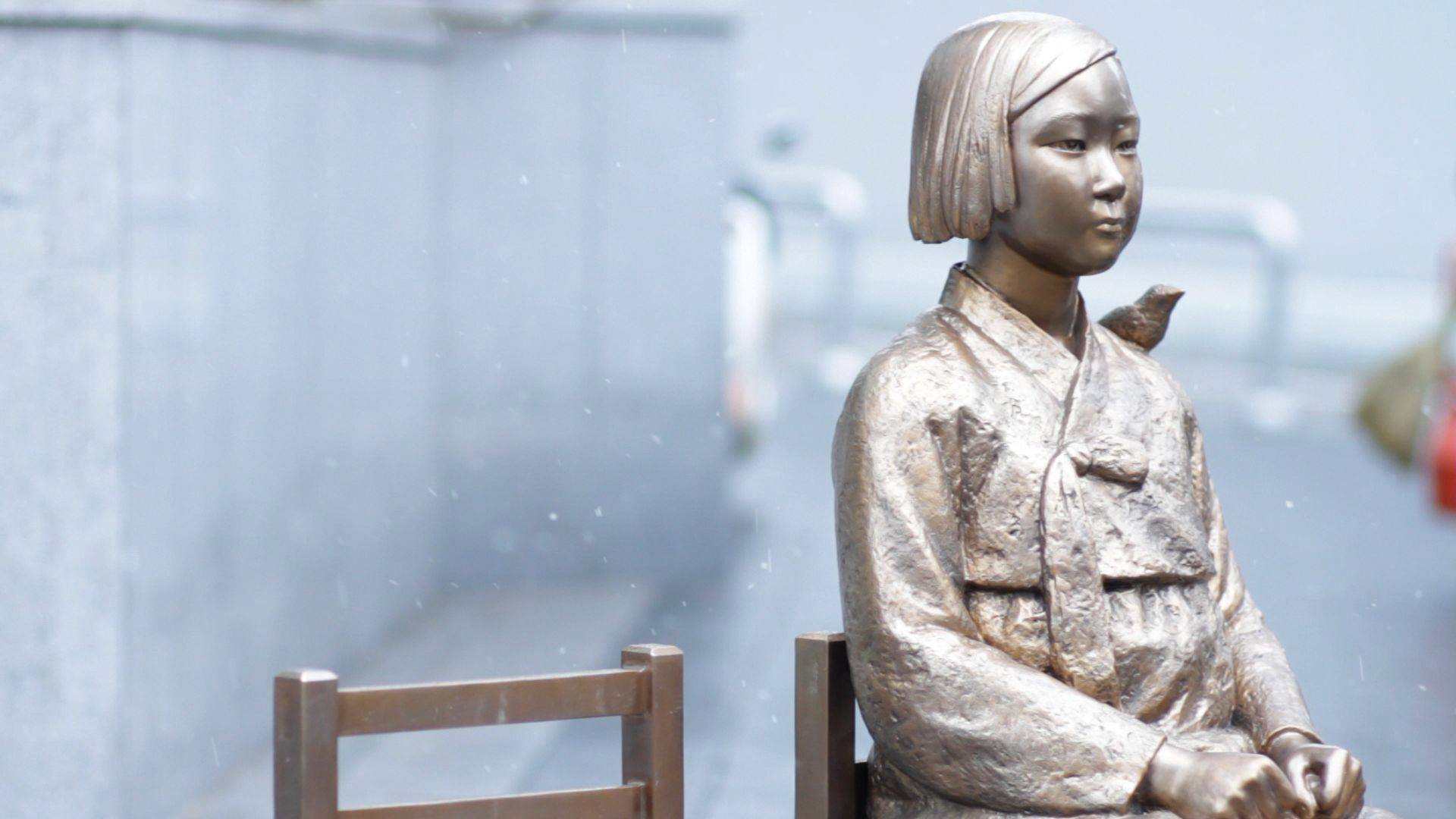 A statue of a Korean girl sitting in a chair with a bird on her shoulder, also known as the Statue of Peace
