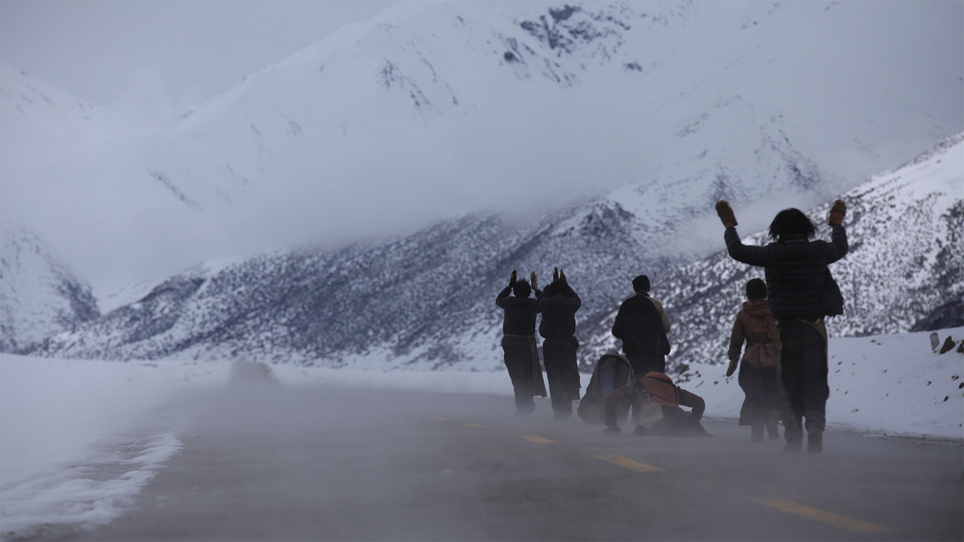 A line of people walk down a snowy mountain roadway in the cold, clapping shoes above their heads