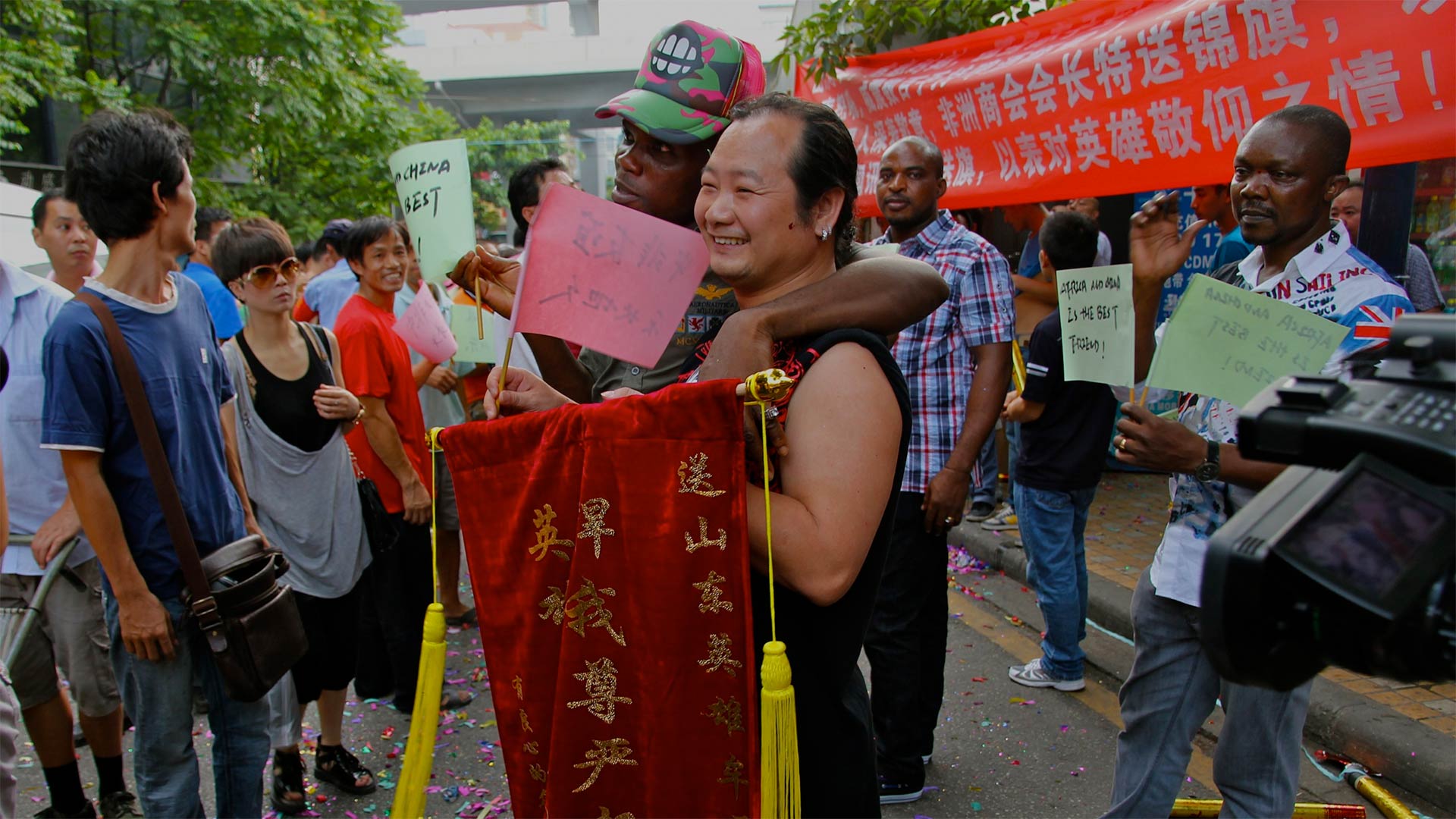 Chinese and African people intermingle and pose for a photo in the street with signs in Chinese and a video camera