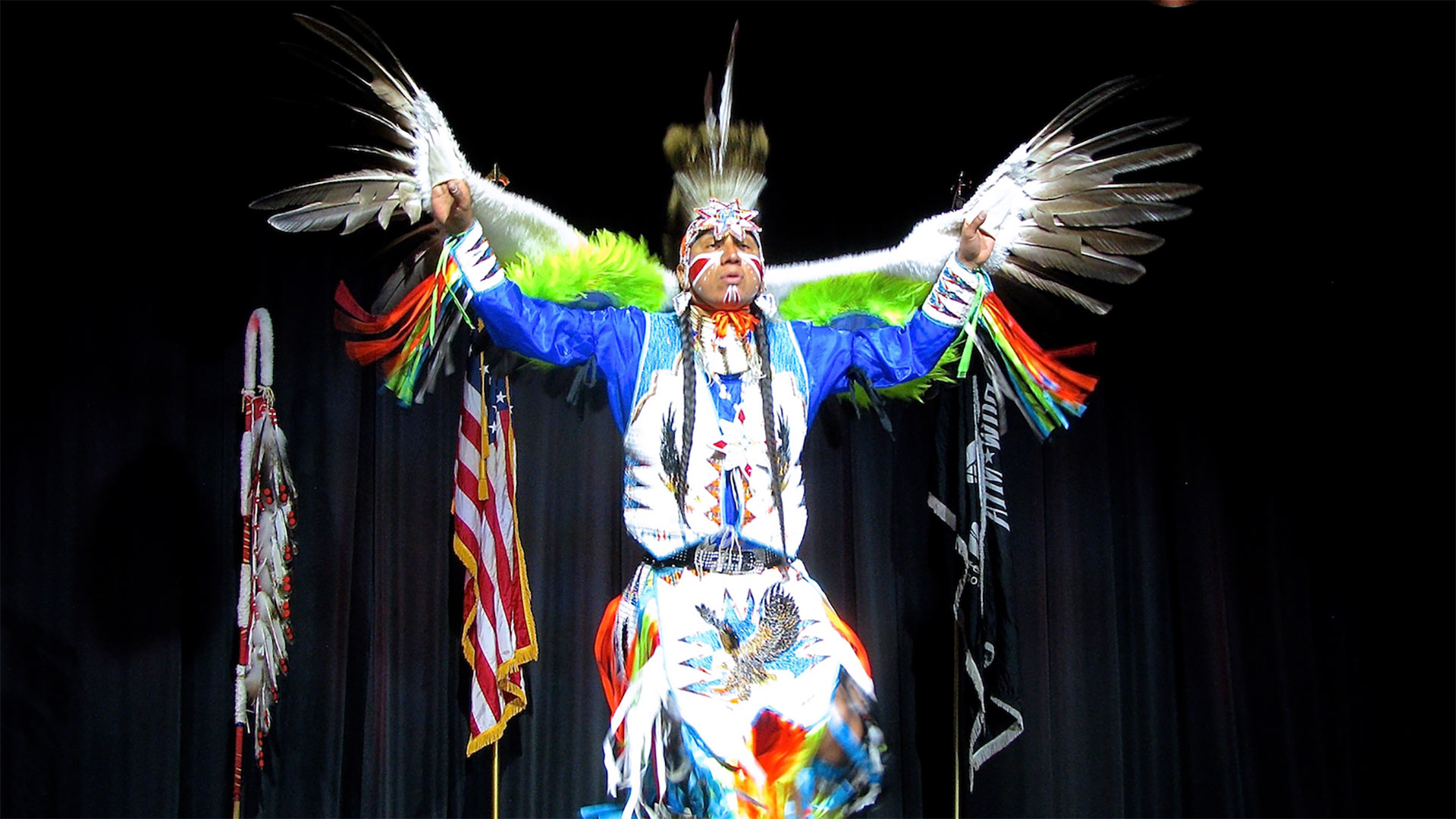 Dancer on stage in colorful dance regalia, with feathers on his arms