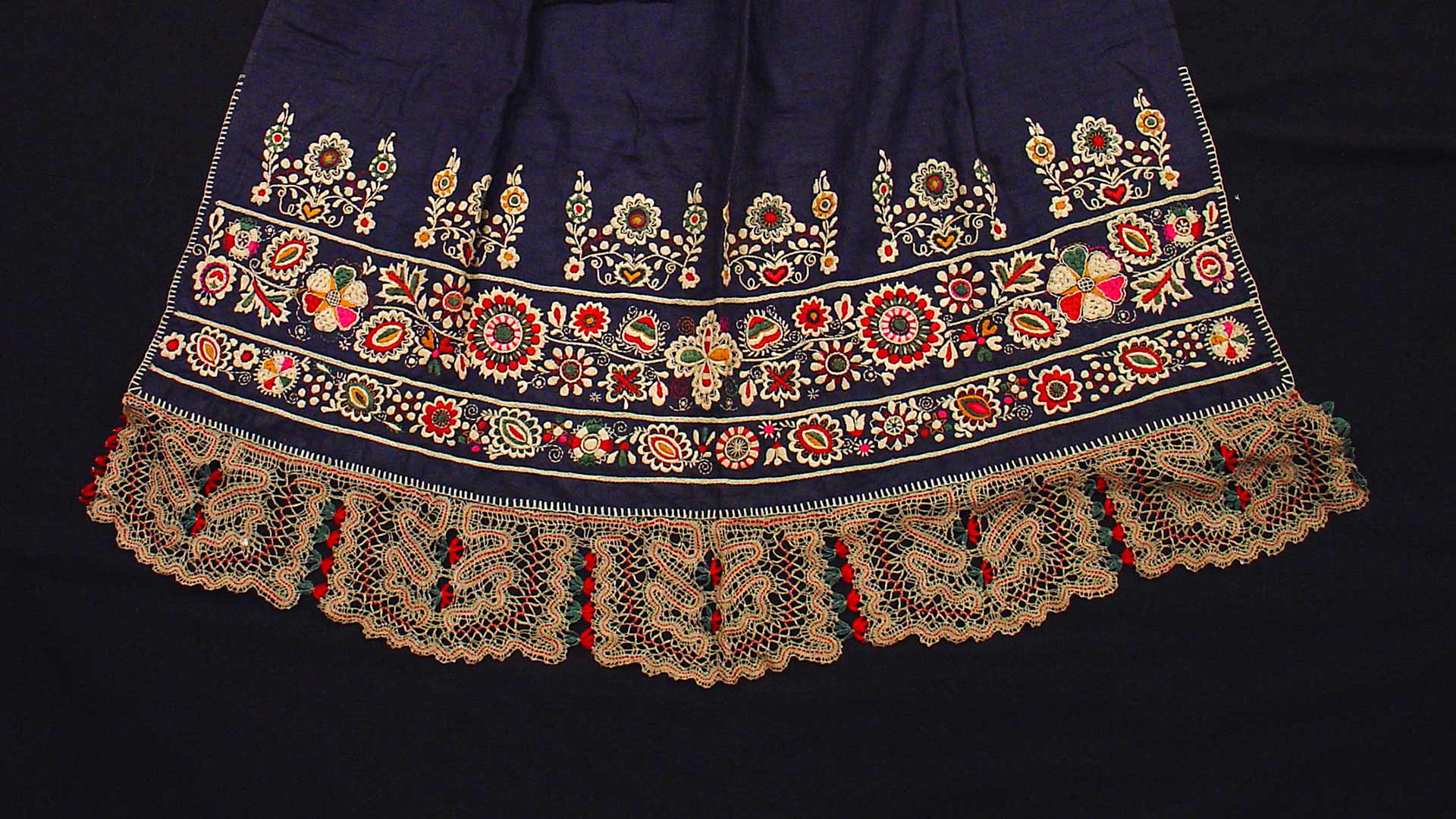 Elaborate embroidery and lace on the bottom of a navy blue skirt. The embroidery and lace are floral patterns of cream, red, green, and pink colors. 