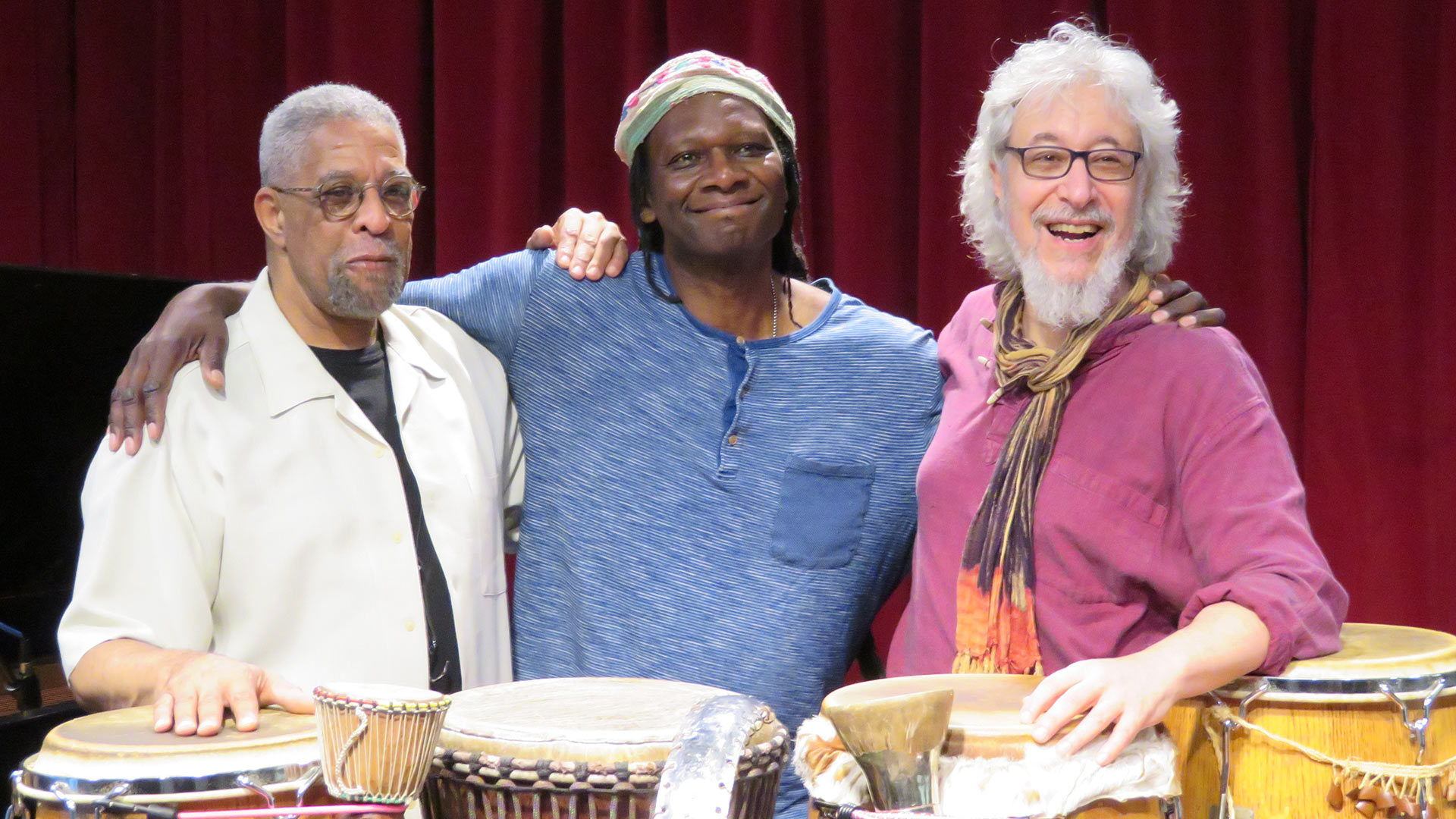 Three musicians smiling and posing for a picture behind four of their drums.