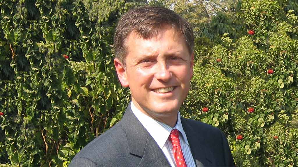 Man in a suit and red tie smiles in the sun in front of lush greenery