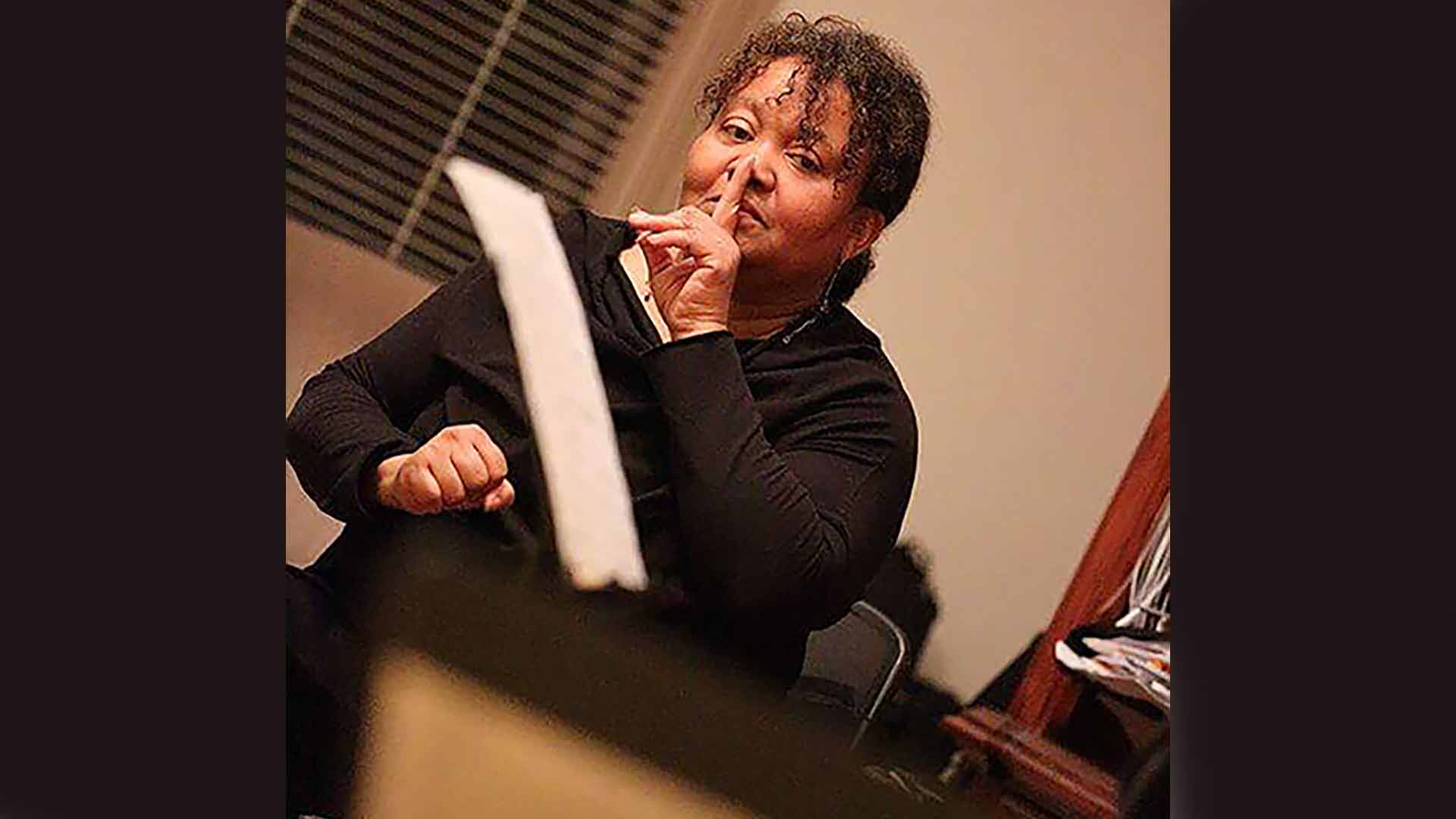 Renee Baker sitting down in chair with a finger pressed upon her lips. 