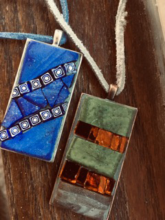 A rectangular, blue mosaic necklace beside another rectangular mosaic--a green one with two orange streaks.